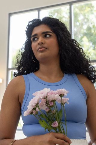 Young woman holding pink chrysanthemum bouquet in blue tank top, looking left by window