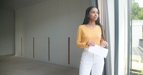 African american woman standing by sliding glass door holding envelope in serene apartment