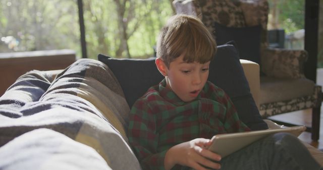 Young boy sitting on a couch using a tablet with a surprised expression. Natural daylight coming through large windows. Can be used in contexts such as technology for kids, education, entertainment at home, or family life. Perfect for articles, blogs, or advertisements focused on children's digital learning, modern parenting, or healthcare and development.