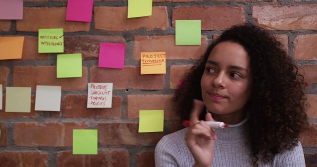 Young biracial professional looking at colorful sticky notes on brick wall. She has curly brown hair, light brown skin, and wears simple top