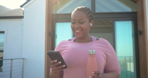 Smiling african american woman checking smartphone holding water bottle on sunlit balcony