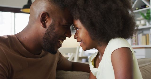 Father and young daughter smiling at each other at home, emphasizing strong family bond and parental love. Ideal for use in campaigns promoting family values, parenting tips, or home life products. This image also works well for articles on fatherhood, child development, and emotional connection.