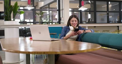 Young African American woman smiling while using phone in coworking lounge with laptop