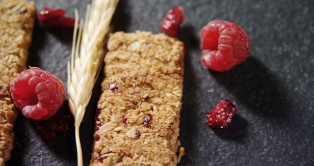 This image depicts a close-up of oat granola bars with fresh raspberries and dried berries scattered around, with a grain stalk also visible. Perfect for use in promotions for healthy snacks, breakfast ideas, nutrition tips, or food blogs highlighting natural and wholesome foods.