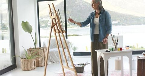 Mature artist painting in bright waterfront studio at wooden easel overlooking hills