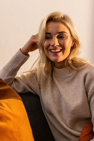 Smiling blonde woman touching hair on cozy sofa with mustard cushion in warm sunlight