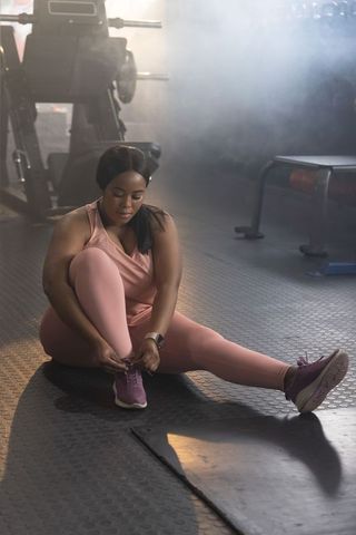 African american woman tying sneaker on gym floor wearing pink activewear and leggings