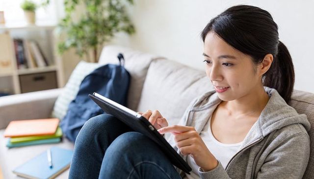Asian woman tapping tablet on sofa studying online learning and remote education at home