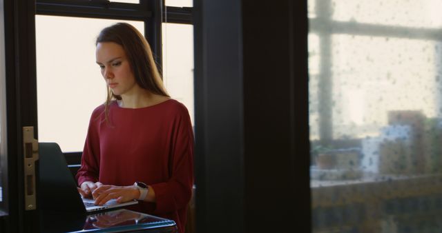 A woman wearing red blouse is standing at her desk while typing on her laptop in a modern office. The large windows in the background create an environment with natural light and emphasize the contemporary interior design. This is perfect for portraying a professional and productive working environment, suitable for business, technology, and corporate culture themes.