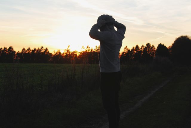 Silhouetted man standing on rural path during sunset, hands on head in contemplative pose. Suitable for illustrating themes of reflection, calmness, decision-making, and connection with nature. Ideal for websites, blogs, or advertisements focused on outdoor activities, mental health, or relaxation.