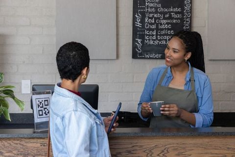 Friendly barista smiling while serving coffee as customer scans qr code at modern cafe counter
