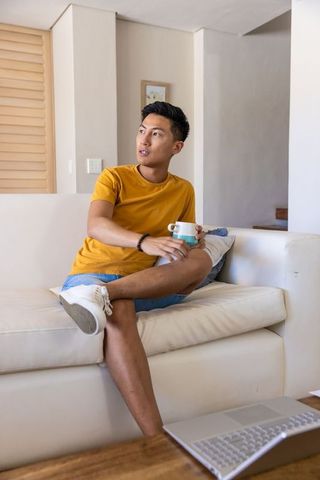 Asian man sitting on white sofa holding teal-and-white mug in contemporary living room