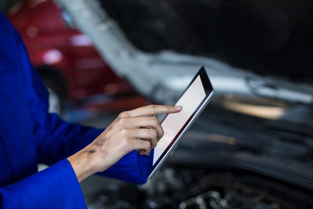 Female mechanic in blue uniform using digital tablet for car maintenance in auto repair garage. Useful for illustrating modern automotive technology, digital tools in car repair, and women in technical professions.