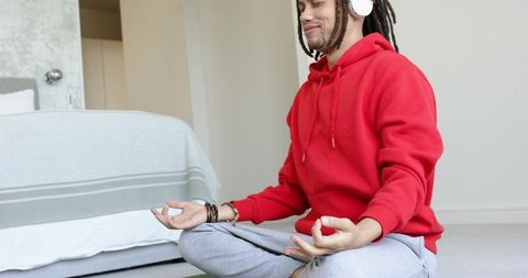 Mid-adult man meditating in minimalist bedroom wearing red hoodie and wireless headphones