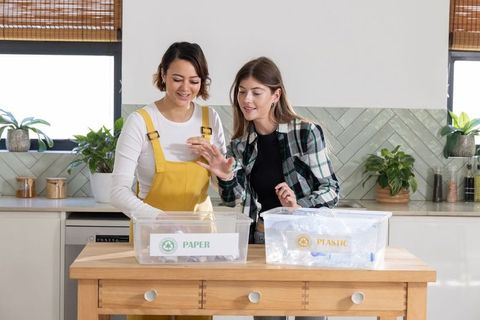Two women sorting recyclables in bright kitchen bins labeled paper and plastic, sustainable living