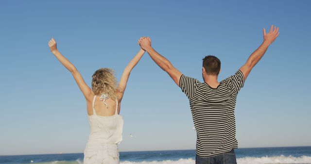 Caucasian couple raising and holding hands and looking at sea at beach. Vacation, summer and lifestyle, unaltered.