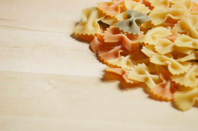 Close-up view of colorful bow tie pasta scattered on a light wooden surface. Ideal for use in food blogs, recipe websites, or culinary articles focusing on Italian cuisine and pasta dishes.