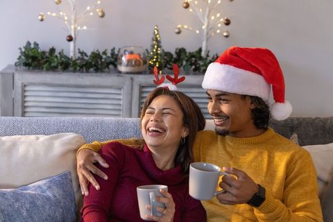 Multicultural couple laughing and holding mugs while relaxing on sofa with christmas hats