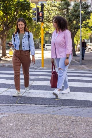 Two women walking across city crosswalk wearing denim jacket and pink shirt urban style
