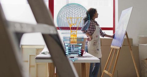 Artist painting at easel in sunlit studio wearing paint-stained apron with mixed-media sculpture