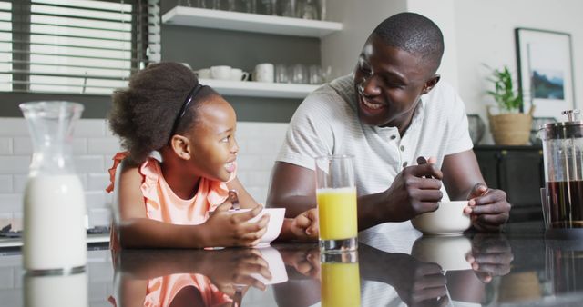 Father and daughter enjoying breakfast together at home, both smiling and engaged. Modern kitchen with sunlight, milk, juice and bowl of cereal. Ideal for concepts of family bonding, morning routines, parenting, and home life activities.