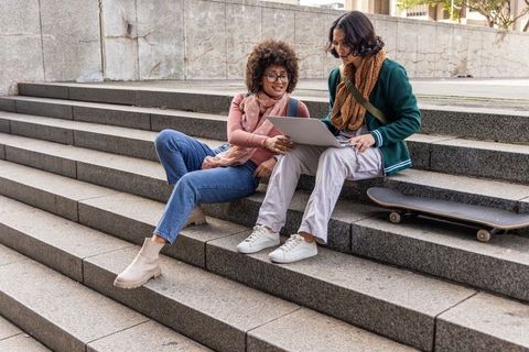 Multicultural female students studying and sharing laptop on urban steps with skateboard