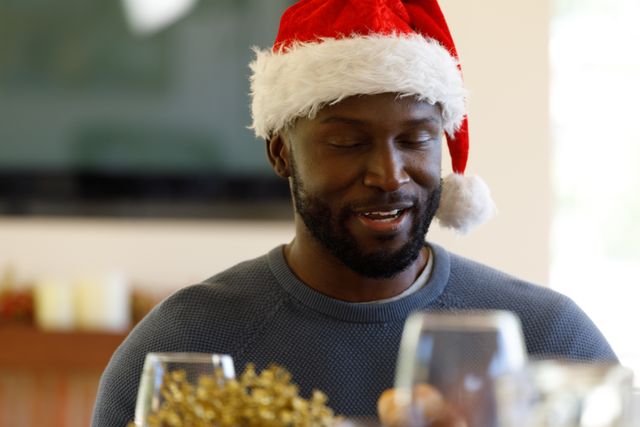 Front view of an african-american man wearing a santa hat while sitting at the christmas dinner table. wine glasses can be seen in the foreground.