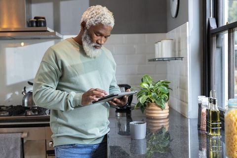 Senior african american man standing in modern kitchen using tablet near coffee mug