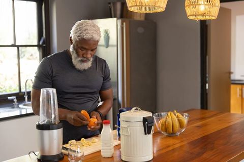 Senior african american man peeling orange and slicing banana preparing healthy smoothie in sunlit k