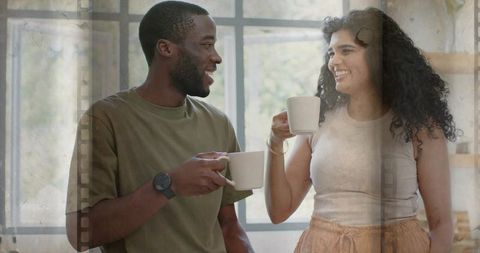 Cozy morning conversation: young couple holding coffee mugs by sunlit grid window