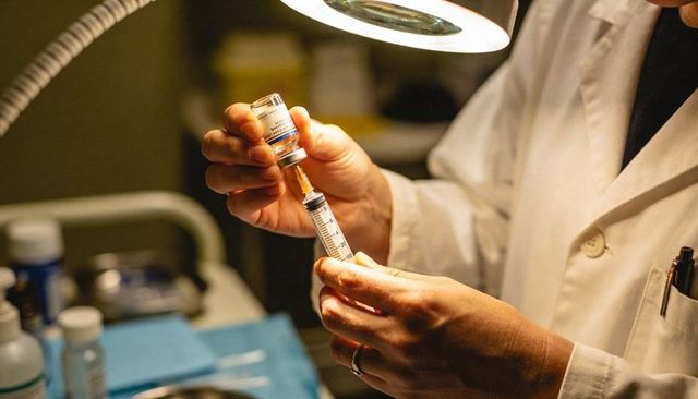 Clinician drawing vaccine into syringe under magnifying lamp wearing white lab coat
