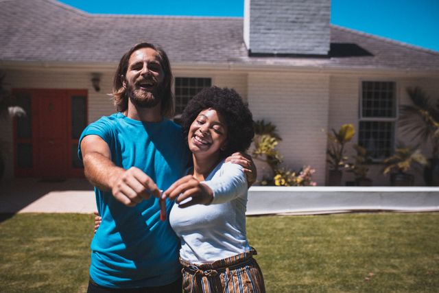 Happy diverse couple standing in their garden, embracing and holding house keys, symbolizing new homeownership. Ideal for use in real estate promotions, home buying advertisements, family lifestyle blogs, and residential property marketing.