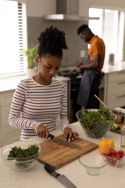 Young African American woman chopping vegetables on kitchen island while man cooks in background. Perfect for illustrating themes of domestic life, healthy eating, and relationship bonding. Ideal for use in lifestyle blogs, cooking websites, and advertisements promoting home and family activities.