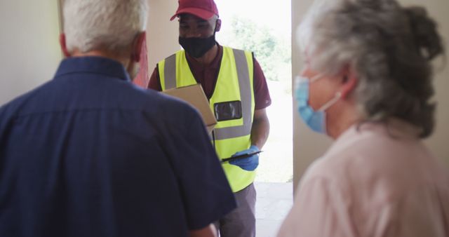 Elderly couple in masks receiving package from delivery person dressed in high-visibility vest and gloves. Suitable for themes on online shopping, logistics, elderly care, pandemic safety, and delivery services.