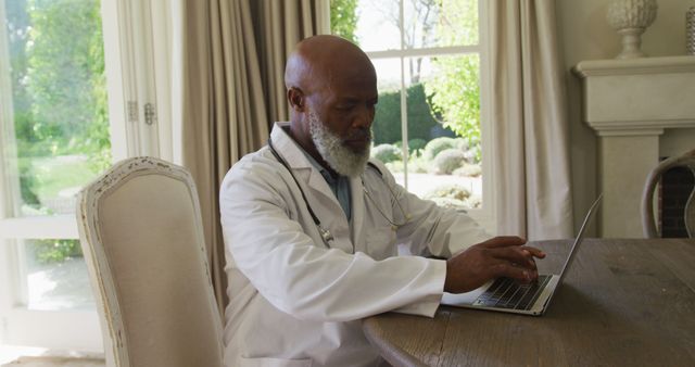 Senior male doctor in white coat working on laptop at home. Ideal for topics related to telemedicine, remote healthcare services, elderly working professionals, modern medical practices, healthcare technology advancements.