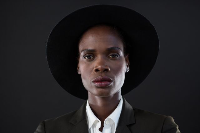 This image depicts an androgynous man wearing a suit and black hat, posing confidently against a grey background. The serious expression and formal attire make it suitable for use in fashion editorials, professional profiles, or advertisements focusing on modern and stylish themes. It can also be used in articles discussing gender identity, fashion trends, or professional attire.