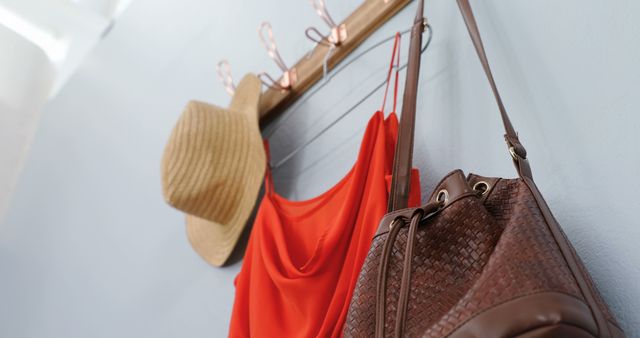 A summer hat, red dress, and woven leather shoulder bag hanging on wall hooks. Perfect for fashion blogs, travel sites, and lifestyle magazines. Provides a rustic yet stylish visual element depicting casual and relaxed summer clothing and accessories. Can be used to illustrate wardrobe organization or home decor ideas.