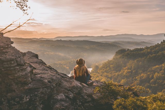 Middle-aged woman sits on rocky cliff while appreciating a stunning sunset over rolling mountains. Perfect for promoting travel destinations, hiking gear, mindfulness retreats, and outdoor adventures. Emphasizes tranquility, solitude, and natural beauty.