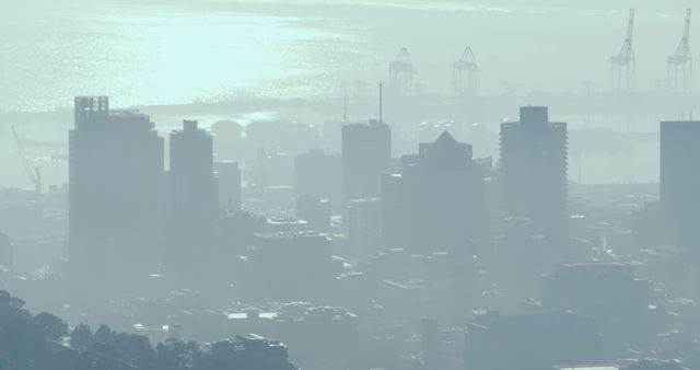 Dense urban skyline veiled by mist with distant industrial cranes complementing sea view. Ideal for urban lifestyle themes, environmental awareness campaigns, and city development projects. Captures essence of modern urban living, transportation infrastructure, and ambiguous beauty of misty mornings.
