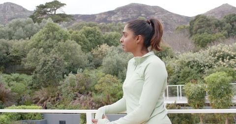 Indian woman on balcony wearing mint athletic top overlooking serene garden and mountain view