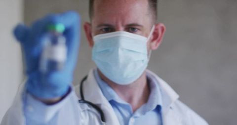 Masked doctor wearing gloves holding vaccine vial in foreground clinical portrait