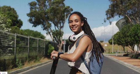 Smiling woman riding electric scooter with backpack and headphones on sunny suburban road