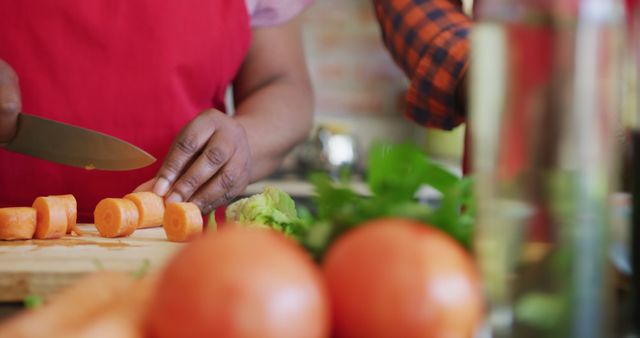 Close-up of a person slicing carrots on a wooden cutting board in a cozy kitchen. The image focuses on healthy food preparation and fresh ingredients. Ideal for use in cooking blogs, recipe websites, healthy eating promotions, and culinary courses.