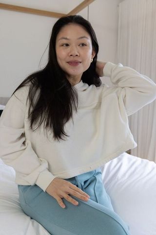 Asian woman sitting on bed wearing cream cropped sweatshirt and blue sweatpants