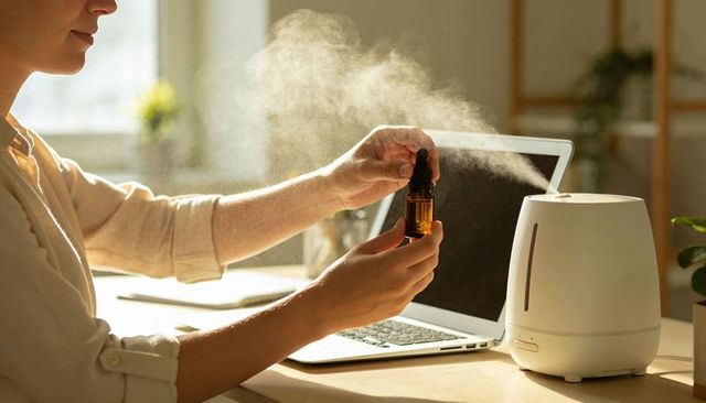 Woman adding essential oil to diffuser at sunlit home office with laptop and mist