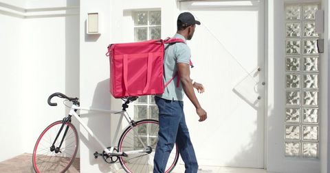 Urban courier walking with bright pink delivery backpack beside white bike at residential entrance