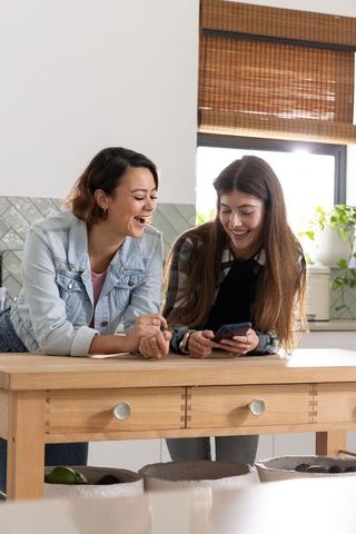 Female friends leaning on kitchen island laughing and sharing smartphone
