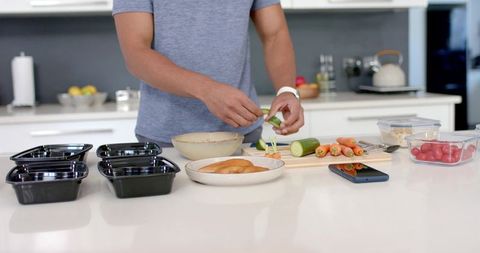 Meal-prepping fresh vegetables in modern kitchen: slicing cucumber, packing black containers
