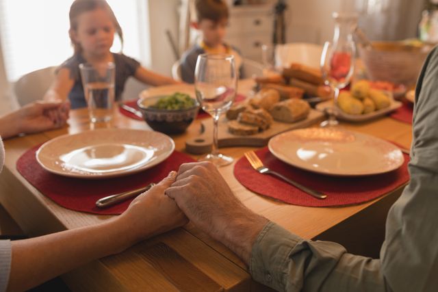 Family holding hands and praying before a meal at home. The table is set with plates, glasses, and various dishes. This image can be used to depict family traditions, gratitude, and togetherness. Ideal for articles or advertisements related to family values, religious practices, or home life.