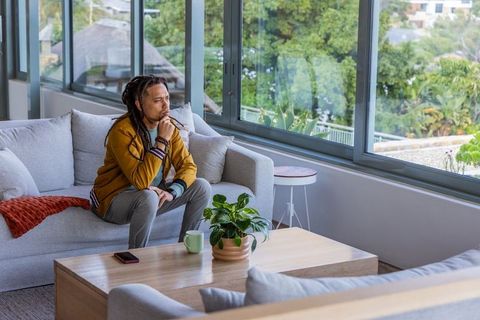 Thirty-something man with dreadlocks sitting on modern grey sofa by large window checking smartphone